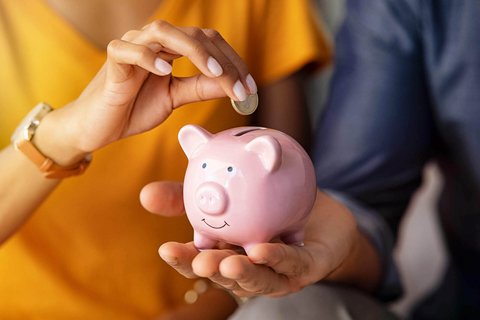 © iStock-1223342144/Ridofranz Ein Mann hält ein rosa Sparschwein, in das eine Frau einen 1 Euro legt.